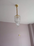 Narrow Clérachic chandelier seen far up, hanging from a cord in a tall room with a lilac wall and a pull cord beside it.