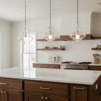 Three Clérachic glass pendants hang above a white kitchen island, with dark wood cabinets and floating shelves in the background.