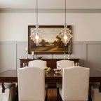 A pair of clear glass pendants from Clérachic hang over a dark wood dining table, in front of a framed landscape painting.