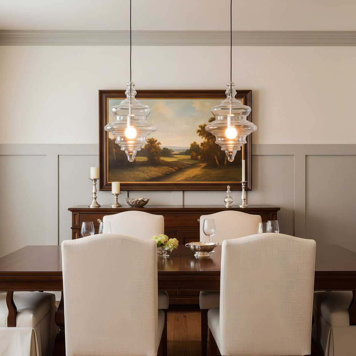A pair of clear glass pendants from Clérachic hang over a dark wood dining table, in front of a framed landscape painting.