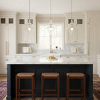 Three clear glass pendants from Clérachic hang in a row over a navy kitchen island with white worktops and woven stools.
