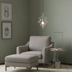 Clérachic clear glass pendant with a lit bulb, hanging over a grey armchair and footstool in a sage green room.