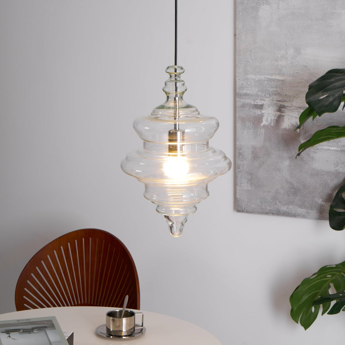 Lit tiered clear glass Clérachic pendant on a black cord, in front of a grey abstract painting and a green plant leaf.
