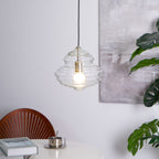 Wide, ribbed clear glass pendant from Clérachic with a lit bulb, hanging above a white table with a book and a small metal cup.