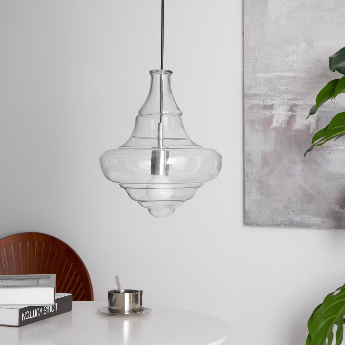 Bell-shaped clear glass pendant from Clérachic with a silver-toned holder and a bulb inside, hanging beside a leafy plant.