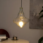 A Clérachic bell-shaped glass pendant lit in a warmer, dimmer room, with a white table and a dark wooden chair back below.