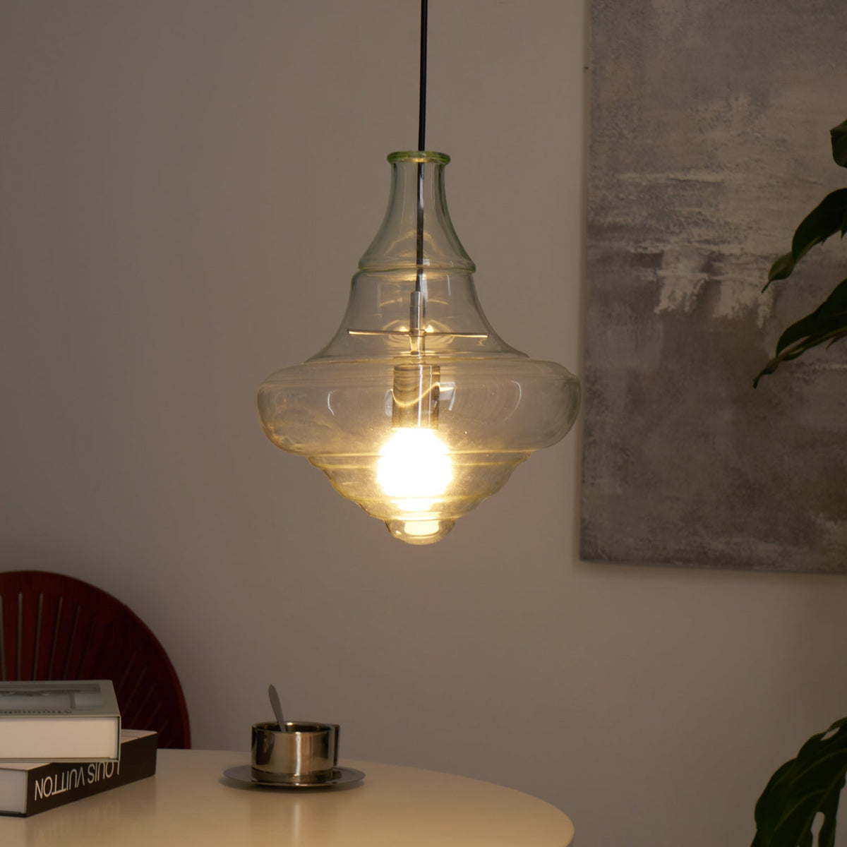 A Clérachic bell-shaped glass pendant lit in a warmer, dimmer room, with a white table and a dark wooden chair back below.
