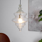 Tiered clear glass Clérachic pendant with a lit bulb and a silver-toned holder, hanging beside a framed grey abstract picture.