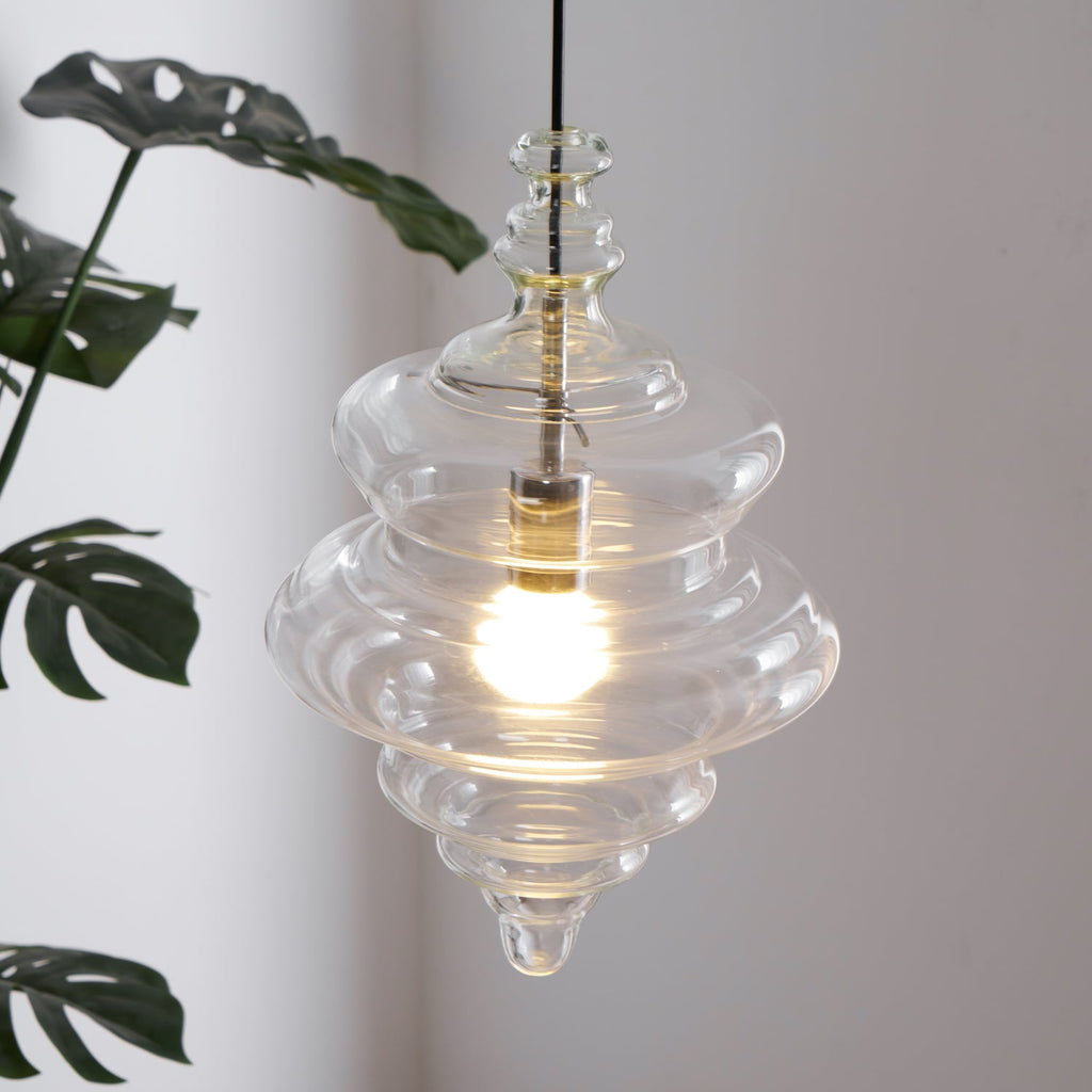Close view of a tiered clear glass pendant from Clérachic, glowing warm in front of large green leaves.