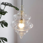 Close view of a tiered clear glass pendant from Clérachic, glowing warm in front of large green leaves.
