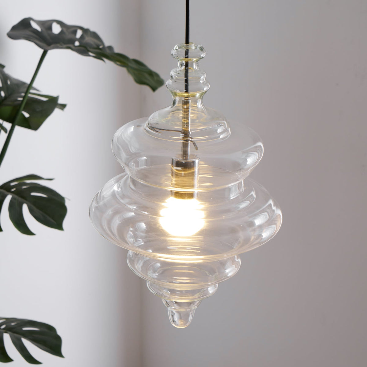 Close view of a tiered clear glass pendant from Clérachic, glowing warm in front of large green leaves.