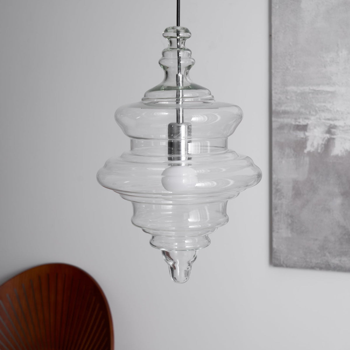 Unlit tiered clear glass Clérachic pendant with a silver-toned holder and a pointed tip, in front of a grey painting and a wooden chair.
