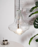 Clear glass Clérachic pendant with a bell-shaped top and a wide base, lit above a table with a metal cup and a green leaf.