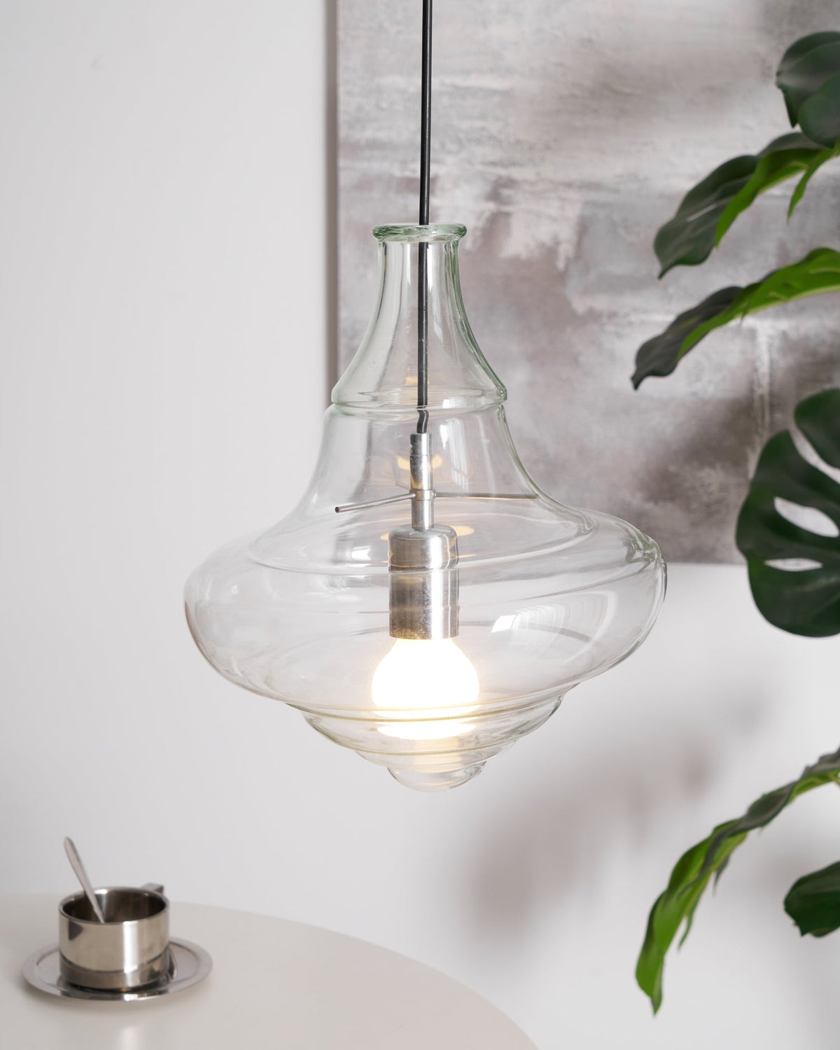 Clear glass Clérachic pendant with a bell-shaped top and a wide base, lit above a table with a metal cup and a green leaf.