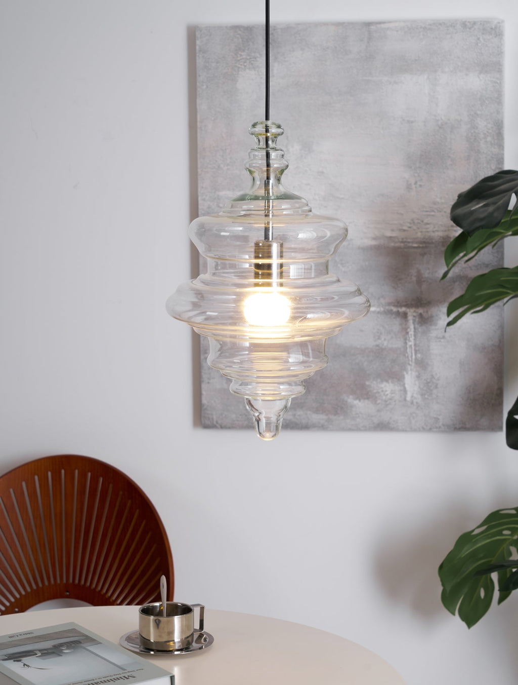 Lit tiered glass pendant from Clérachic hangs in front of a grey painting, with a table, book and plant in the lower part of the frame.