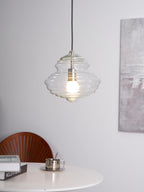 Small ribbed clear glass Clérachic pendant glowing in a white room, with a table and a wooden chair back below.