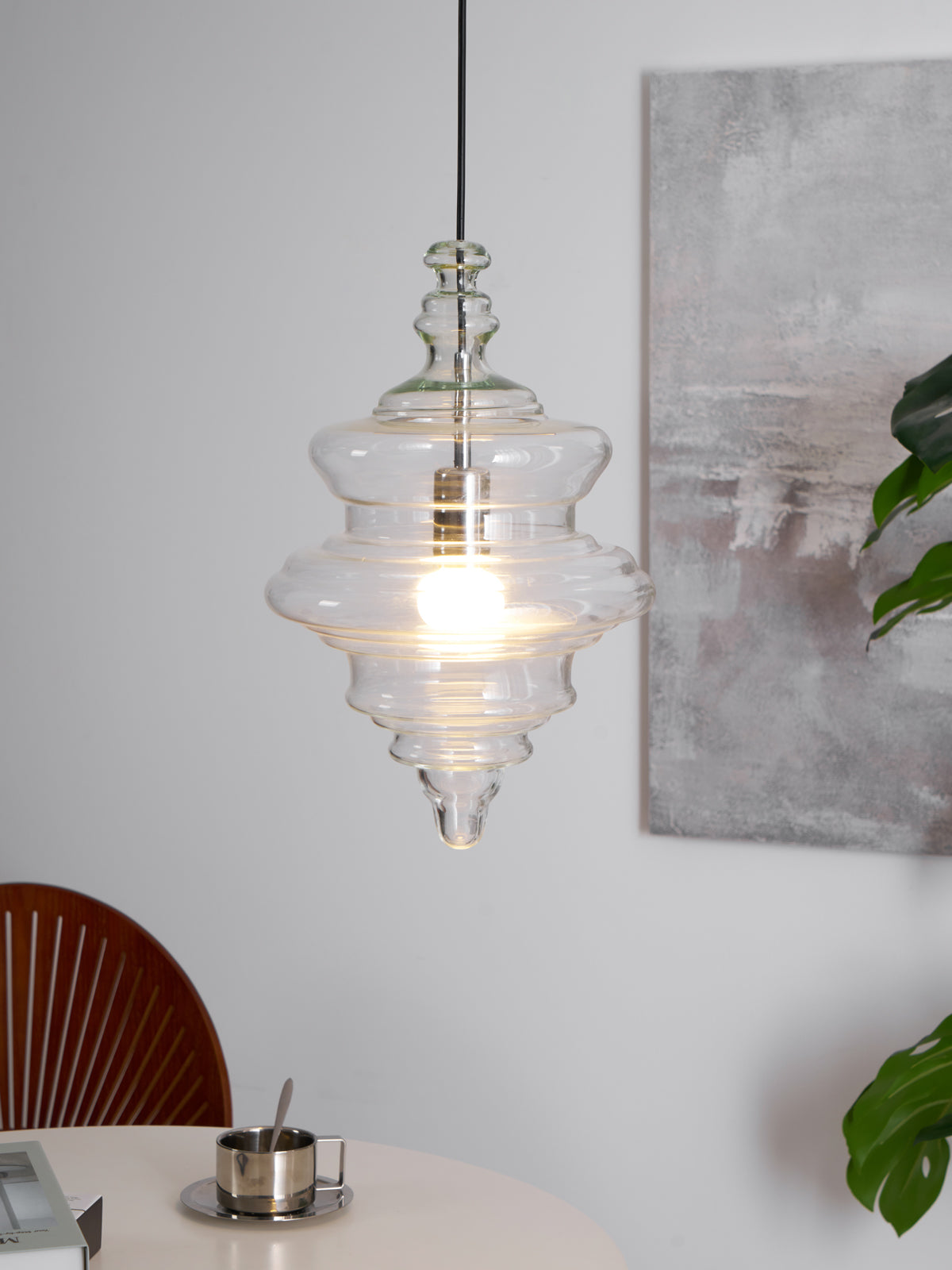 A tiered Clérachic glass pendant lights up in front of a grey painting, with a plant leaf at the right edge of the frame.