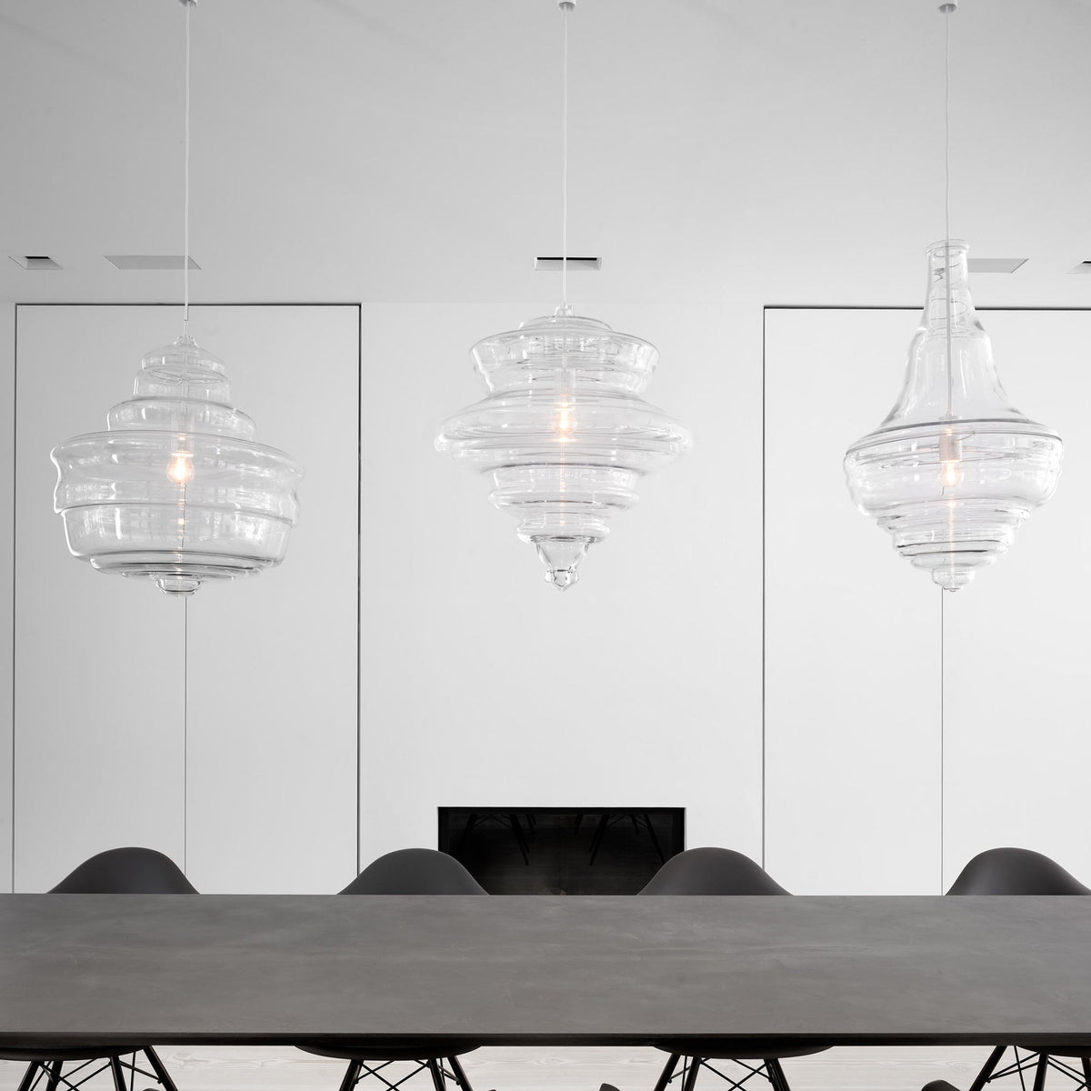 Three clear glass Clérachic pendants in different shapes hang above a long grey table with black chairs, in a white room.