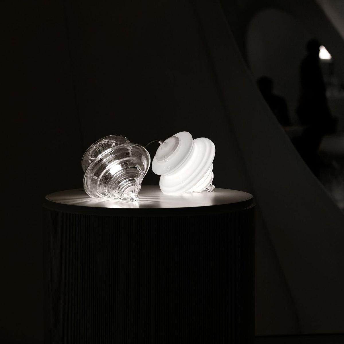 A clear glass shade and a white opal one sit side by side on a dark round table, both lit, in a Clérachic still life.