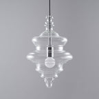 Tiered clear glass Clérachic pendant with a bulb inside, on a mid-grey backdrop with a black cord above.