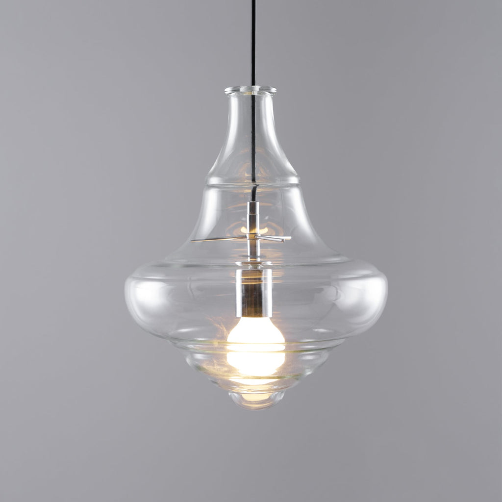 Lit bell-shaped clear glass pendant from Clérachic with a wide, curved base, on a grey backdrop.