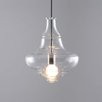 Lit bell-shaped clear glass pendant from Clérachic with a wide, curved base, on a grey backdrop.