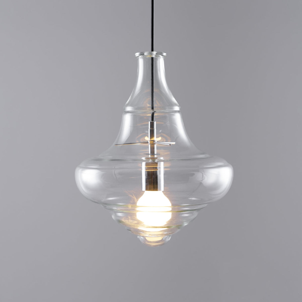 Lit bell-shaped clear glass pendant from Clérachic with a wide, curved base, on a grey backdrop.