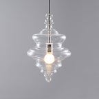Tiered clear glass pendant from Clérachic lit warm on a grey backdrop, with a small finial at the top and a pointed tip.