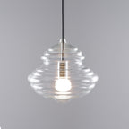 Wide ribbed clear glass Clérachic pendant, with a black cord and a softly lit bulb, on a grey backdrop.