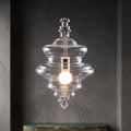 Lit tiered clear glass pendant from Clérachic in a dark textured niche, above a green marble shelf.