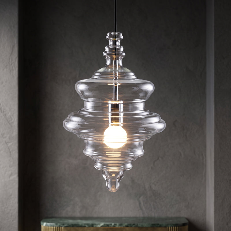 Lit tiered clear glass pendant from Clérachic in a dark textured niche, above a green marble shelf.