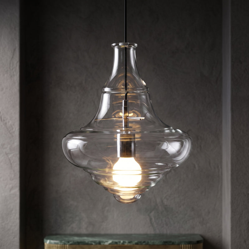A Clérachic bell-shaped clear glass pendant with a lit bulb, hanging in a dark textured alcove above a marble shelf.