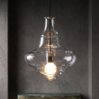 A Clérachic bell-shaped clear glass pendant with a lit bulb, hanging in a dark textured alcove above a marble shelf.
