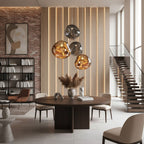 A cluster of Clérachic glass pendants in gold, grey and copper tones hangs above a round dark table in a tall room with brick and timber walls.