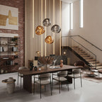 Four Clérachic glass pendants, in gold and smoke tones, hang over a long walnut table in a double-height room with a staircase.
