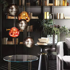 A group of Clérachic glass pendants in copper, gold and smoke tones in front of dark shelves of books, over a glass side table.