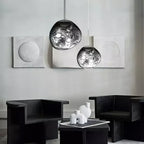 Two Clérachic silver mirror-glass pendants hang in front of grey plaster panels, above two black armchairs and a low table.