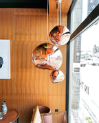 Three Clérachic rose-copper glass pendants of different sizes hang beside a wood-panelled wall and a tall window.