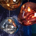 Close view of three Clérachic glass pendants in gold, copper-red and silver against a deep blue backdrop, lit from inside.