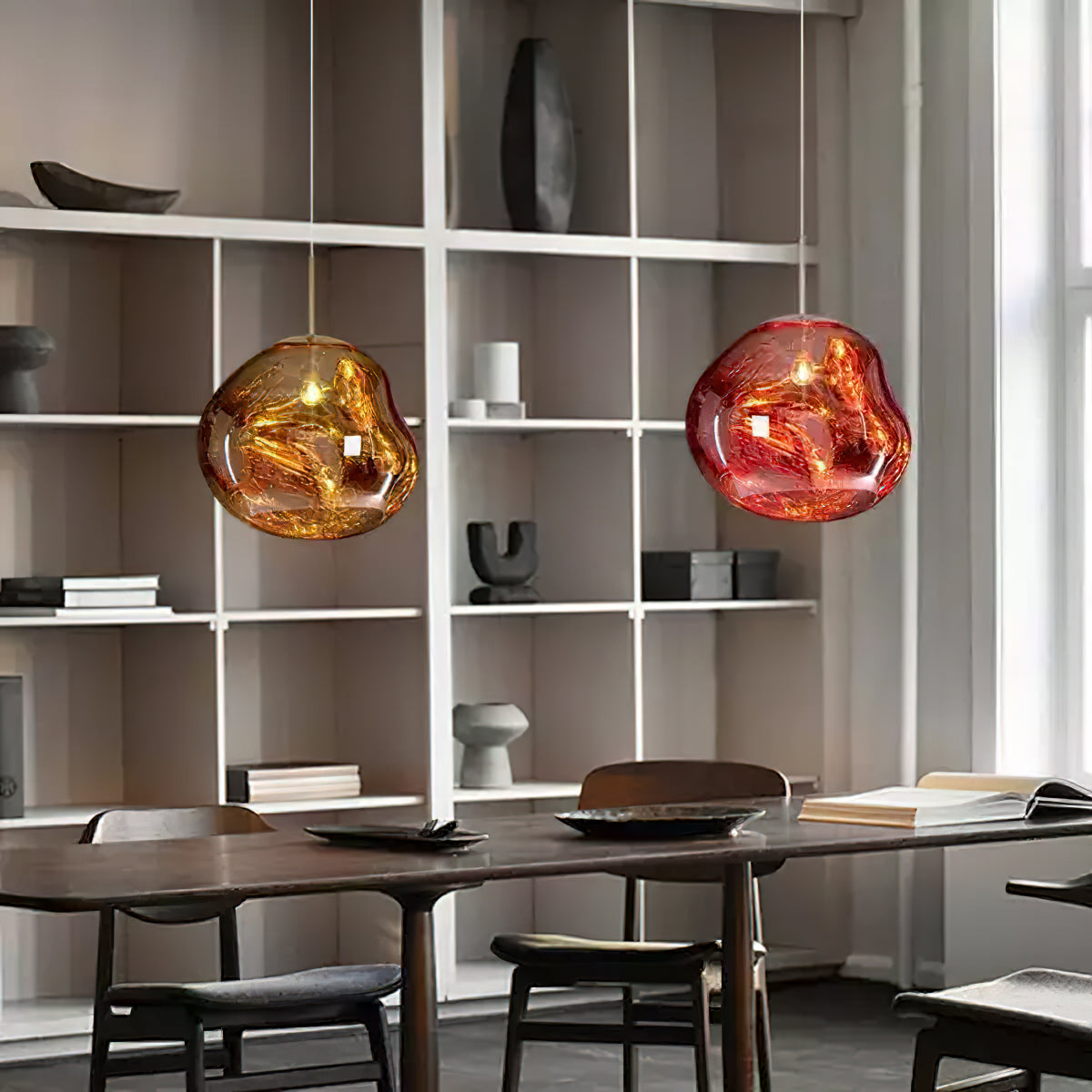 A gold and a copper-red Clérachic pendant hang side by side before white shelving, above a wooden table with chairs.