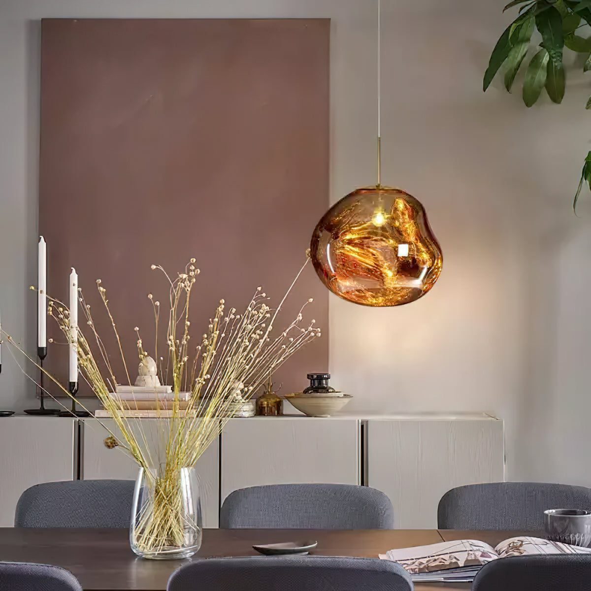 A gold Clérachic glass pendant by a dusty-pink wall, with a vase of dried grass and a pale sideboard with blue chairs.
