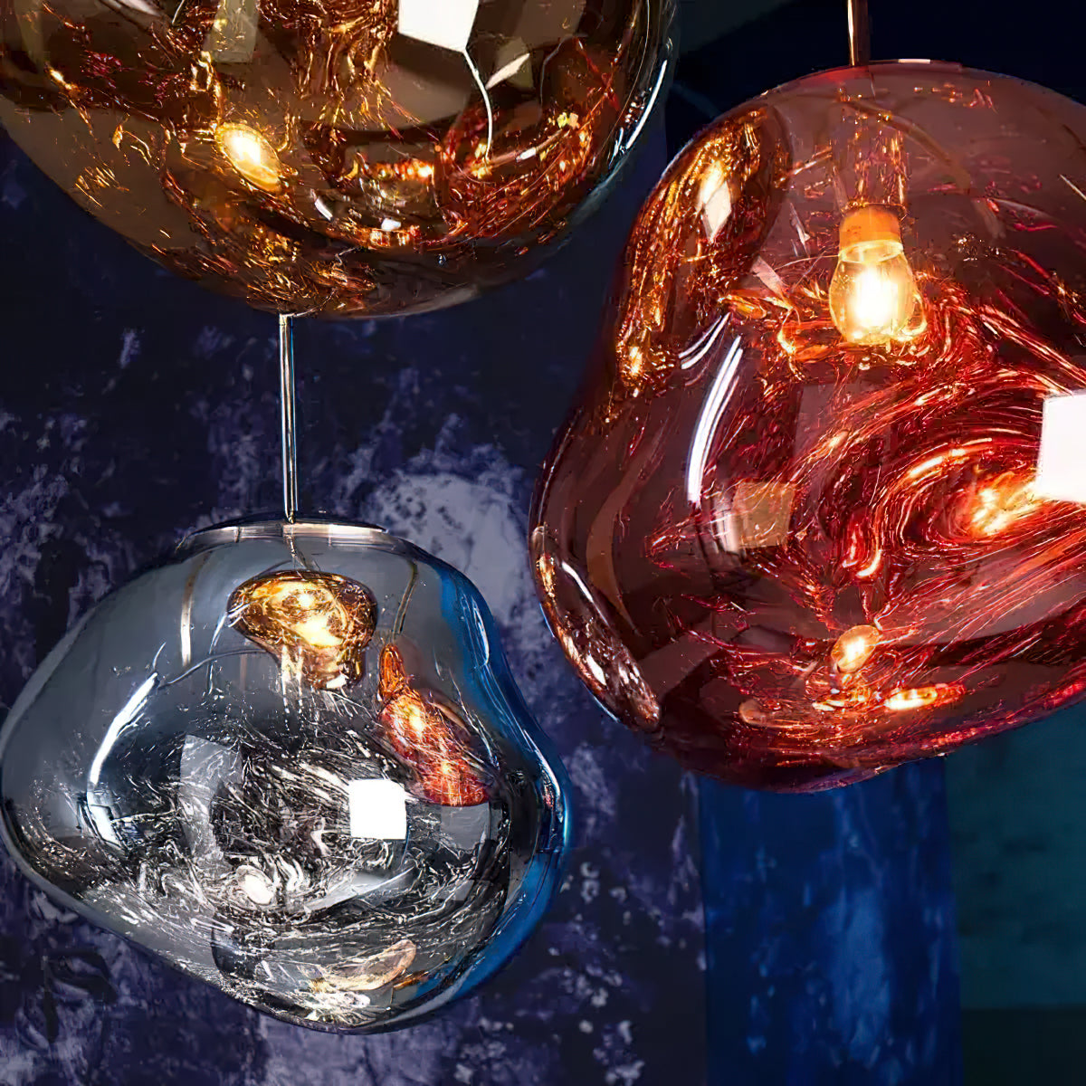 A Clérachic glass pendant in copper-red, a gold one and a silver one on a deep blue backdrop, shown from below with the bulbs lit.