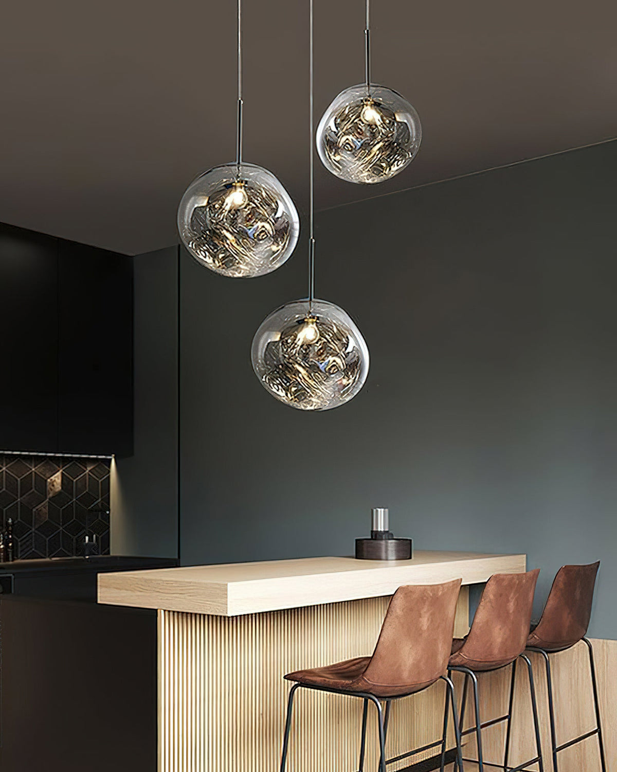 Three Clérachic silver glass pendants above a kitchen bar with leather stools, against a dark green-grey wall.