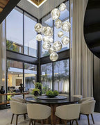 A big group of Clérachic clear silver glass pendants hangs in a glass-walled room, with a round table and cream chairs below.