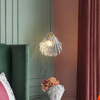 Ribbed clear glass shell pendant from Clérachic glowing against a sage-green wall, between a green velvet chair and a plum curtain.