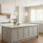 Two Clérachic glass shell pendants hang over a pale kitchen island with a white stone top, in front of a bright window.