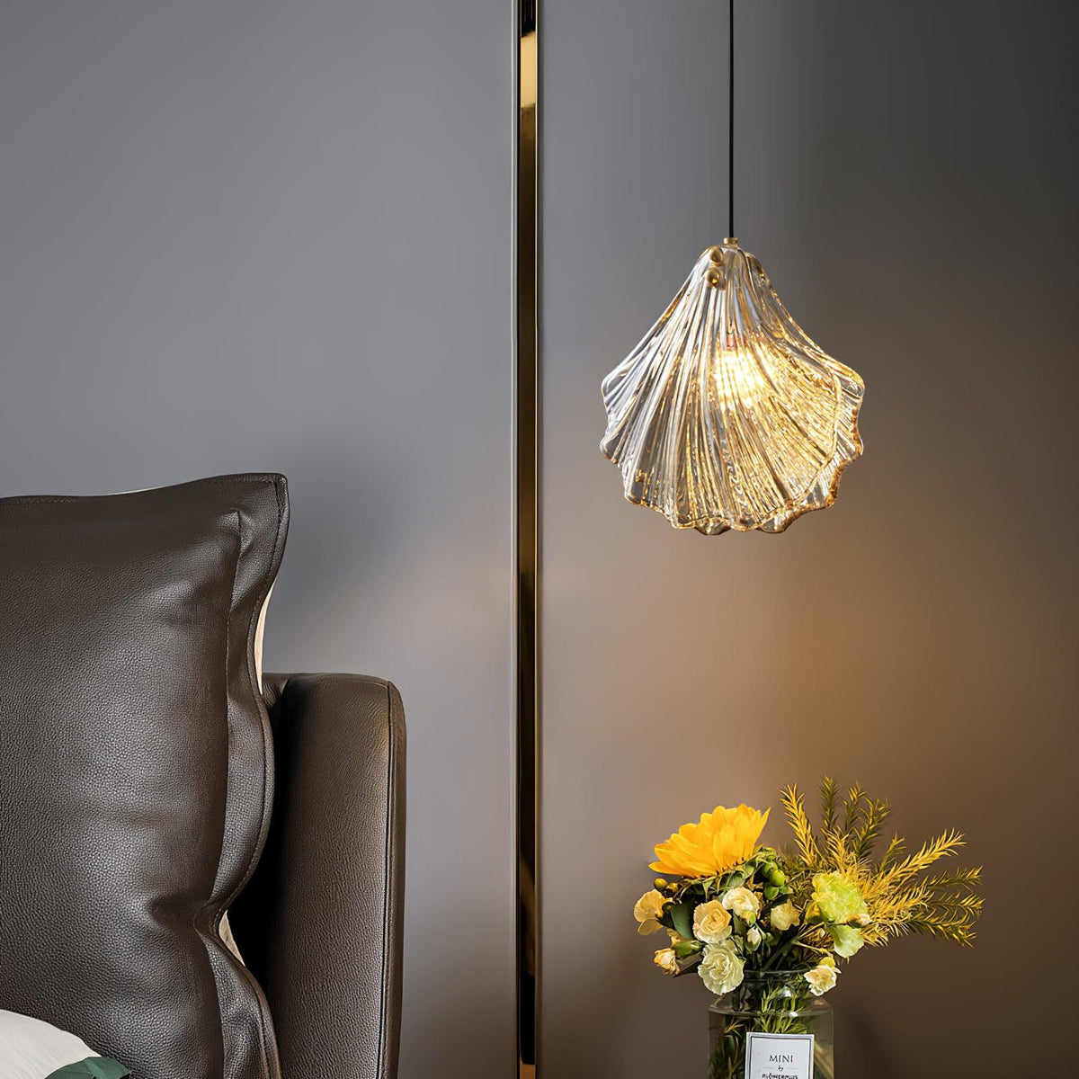 A cognac-toned Clérachic shell pendant lights a grey wall beside a brass trim strip, with a bunch of yellow flowers below.