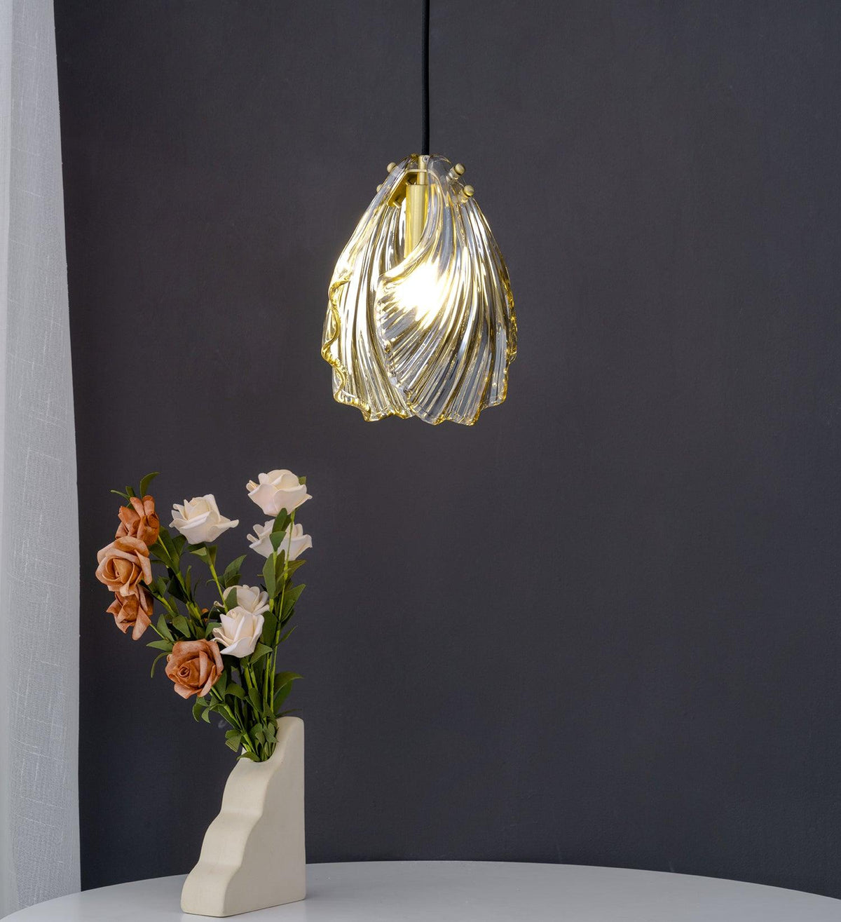 A Clérachic pendant in twisted, spiral-ribbed glass with a brass top hangs beside a vase of roses against a dark wall.