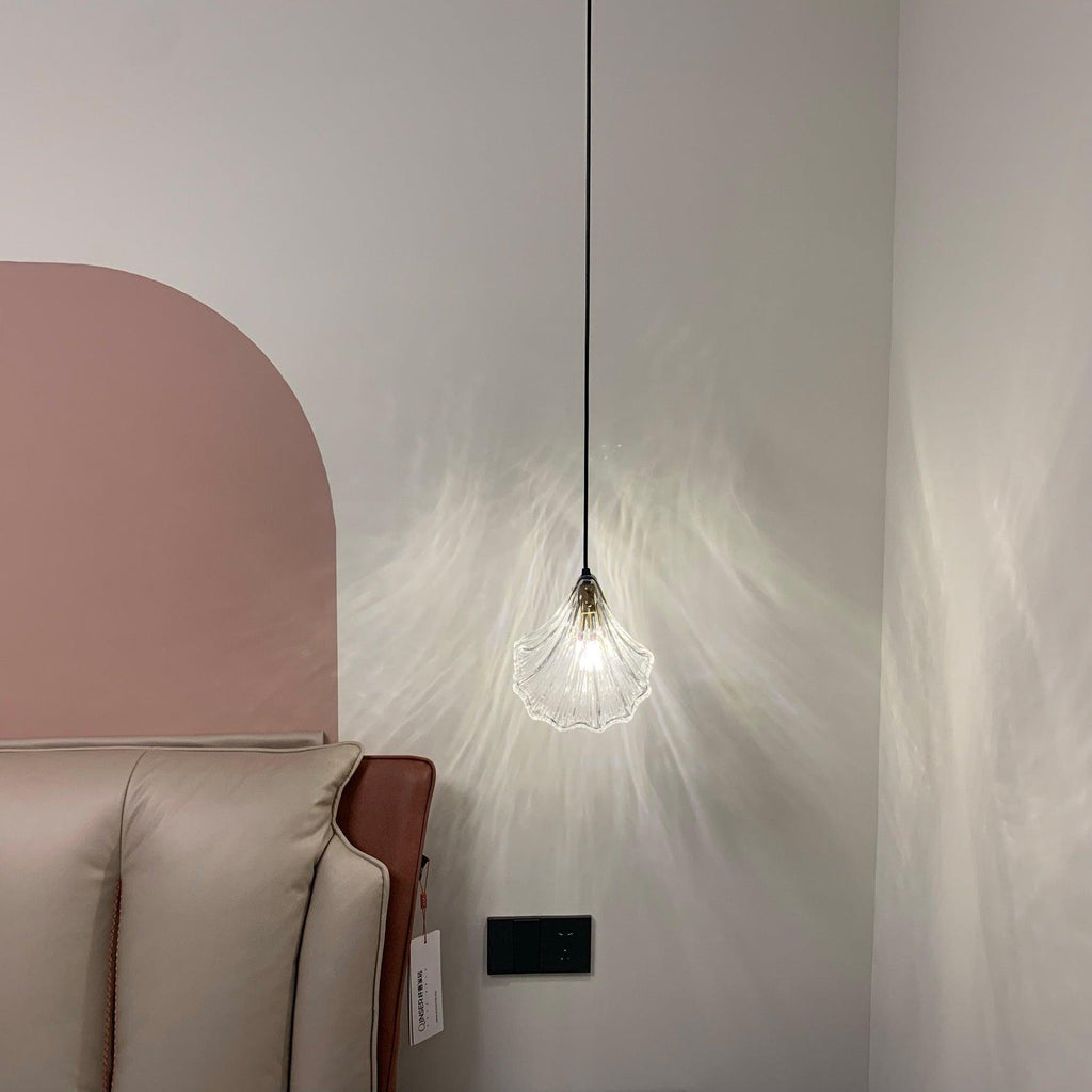 A small lit Clérachic shell pendant on a thin black cord throws a web of light on a pale wall beside a pink arch.