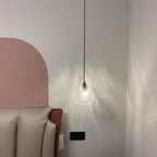 A small lit Clérachic shell pendant on a thin black cord throws a web of light on a pale wall beside a pink arch.